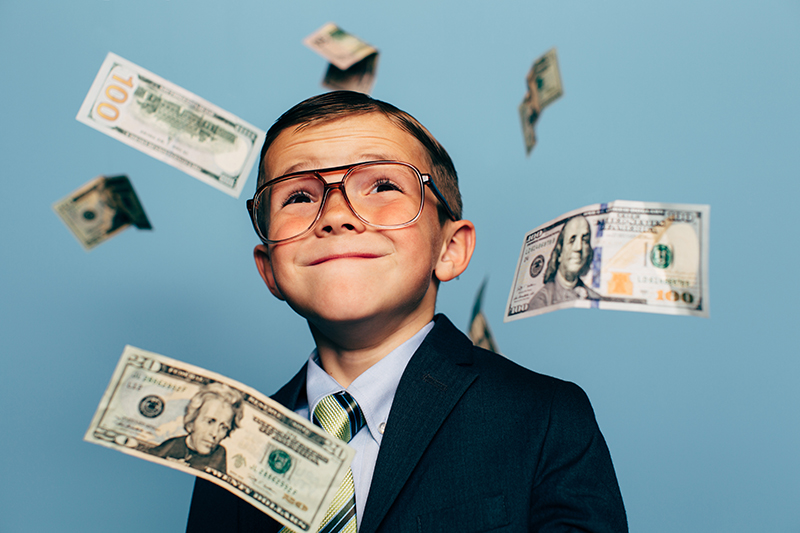 A young boy accountant wearing glasses and suit watches U.S. currency while more falls from above. He is smiling and ready to do your taxes for the IRS and make your tax refund much more money.